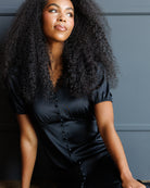 A woman with voluminous dark curly hair wearing a black satin short-sleeve top with a buttoned front, posing against a dark gray paneled wall.