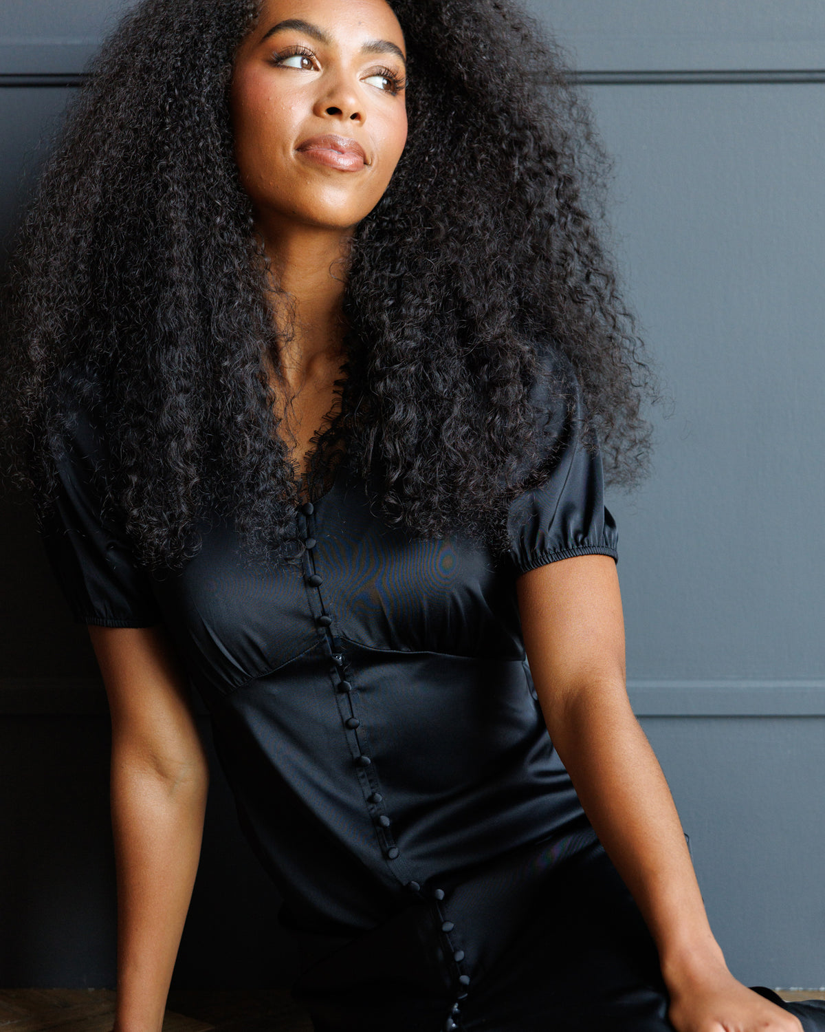 A woman with voluminous dark curly hair wearing a black satin short-sleeve top with a buttoned front, posing against a dark gray paneled wall.