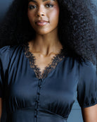 A close-up portrait of a woman wearing a dark navy satin blouse with a black lace V-neck trim and a button-down front.