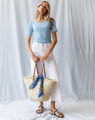 Woman holding a straw bag with a blue scarf, wearing a blue patterned top and white skirt, against a white background.
