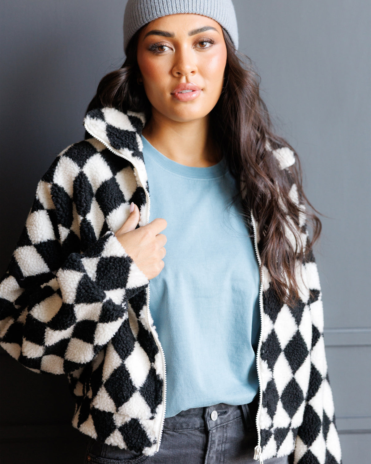 A woman wearing a black and white diamond-patterned sherpa zip-up jacket over a light blue t-shirt and a gray beanie, posing against a dark gray wall.