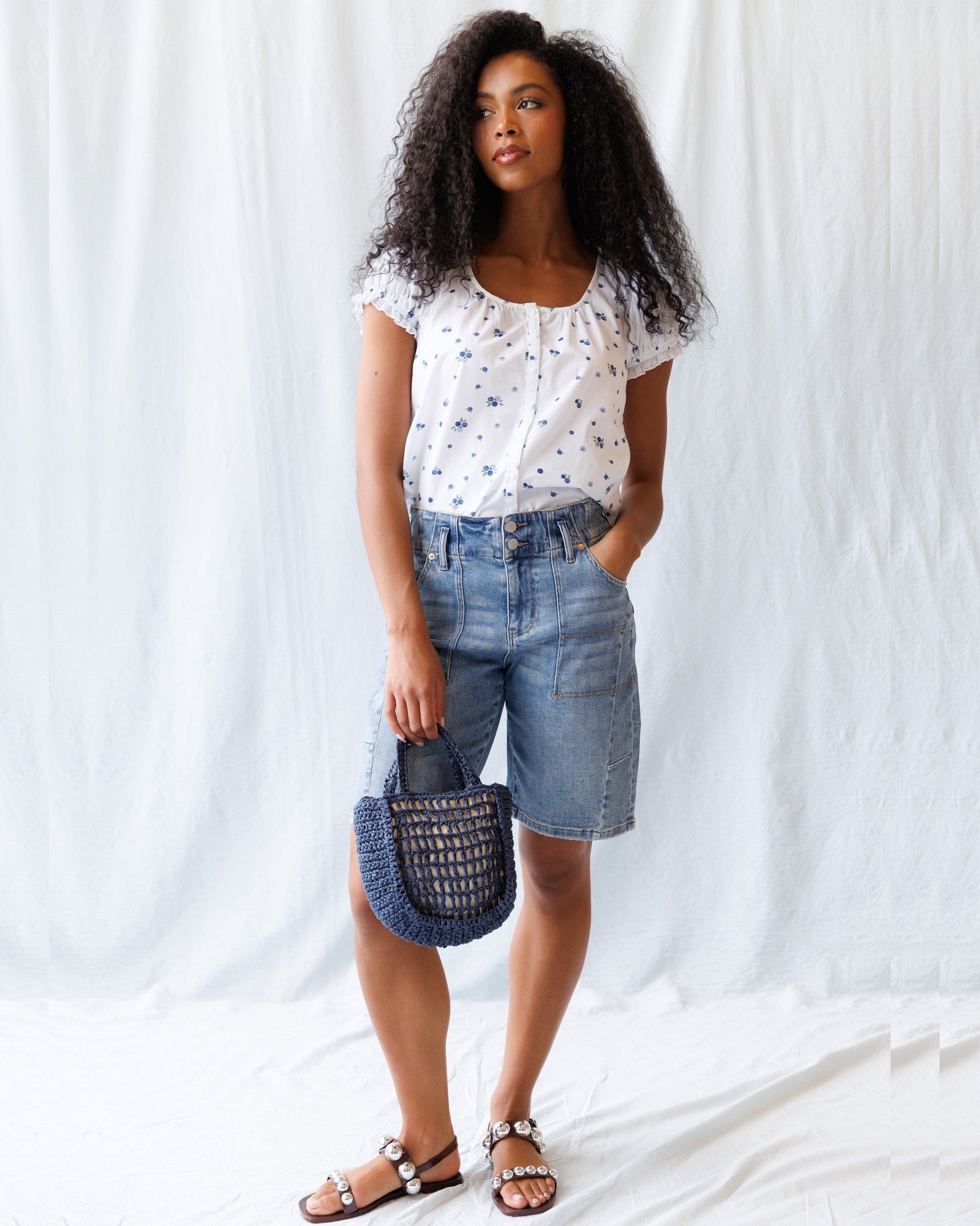 Woman wearing a white floral top and denim shorts, holding a blue bag against a white background