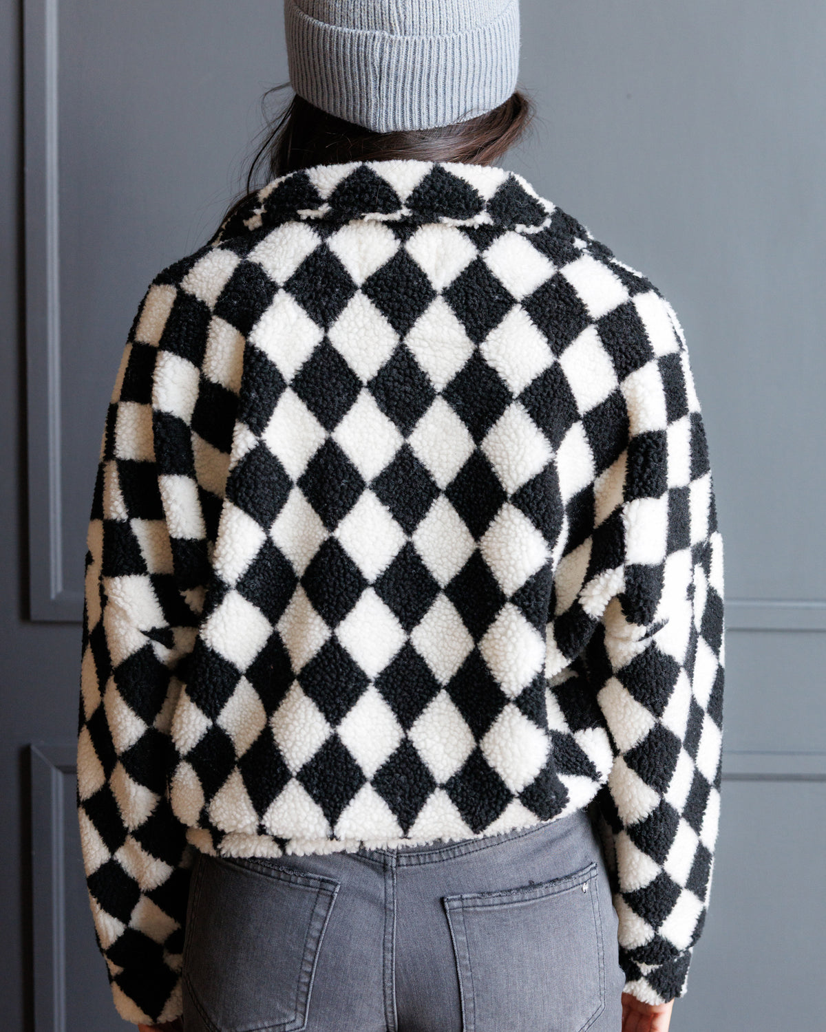 A person shown from the back wearing a black and off-white harlequin diamond-patterned sherpa fleece jacket, paired with gray jeans and a light gray beanie, standing against a dark gray paneled door.