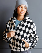A woman wearing a black and white diamond-patterned sherpa fleece zip-up jacket, a light gray beanie, and a blue t-shirt, posing against a dark gray wall.