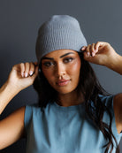 A woman wearing a light gray ribbed knit beanie and a dusty blue sleeveless top, posing with her hands on the sides of the beanie against a dark gray background.