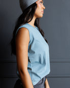 A woman in profile wearing a light blue sleeveless top and a gray knit beanie, standing against a dark gray paneled wall.