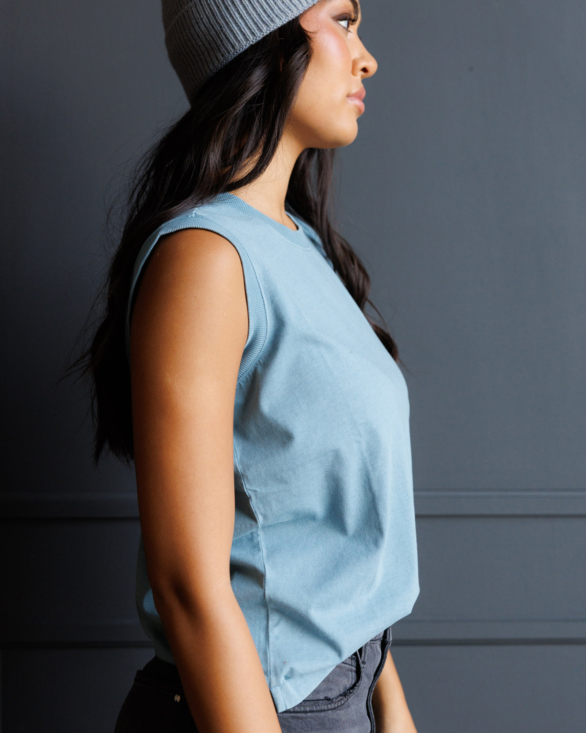 A woman in profile wearing a light blue sleeveless top and a gray knit beanie, standing against a dark gray paneled wall.