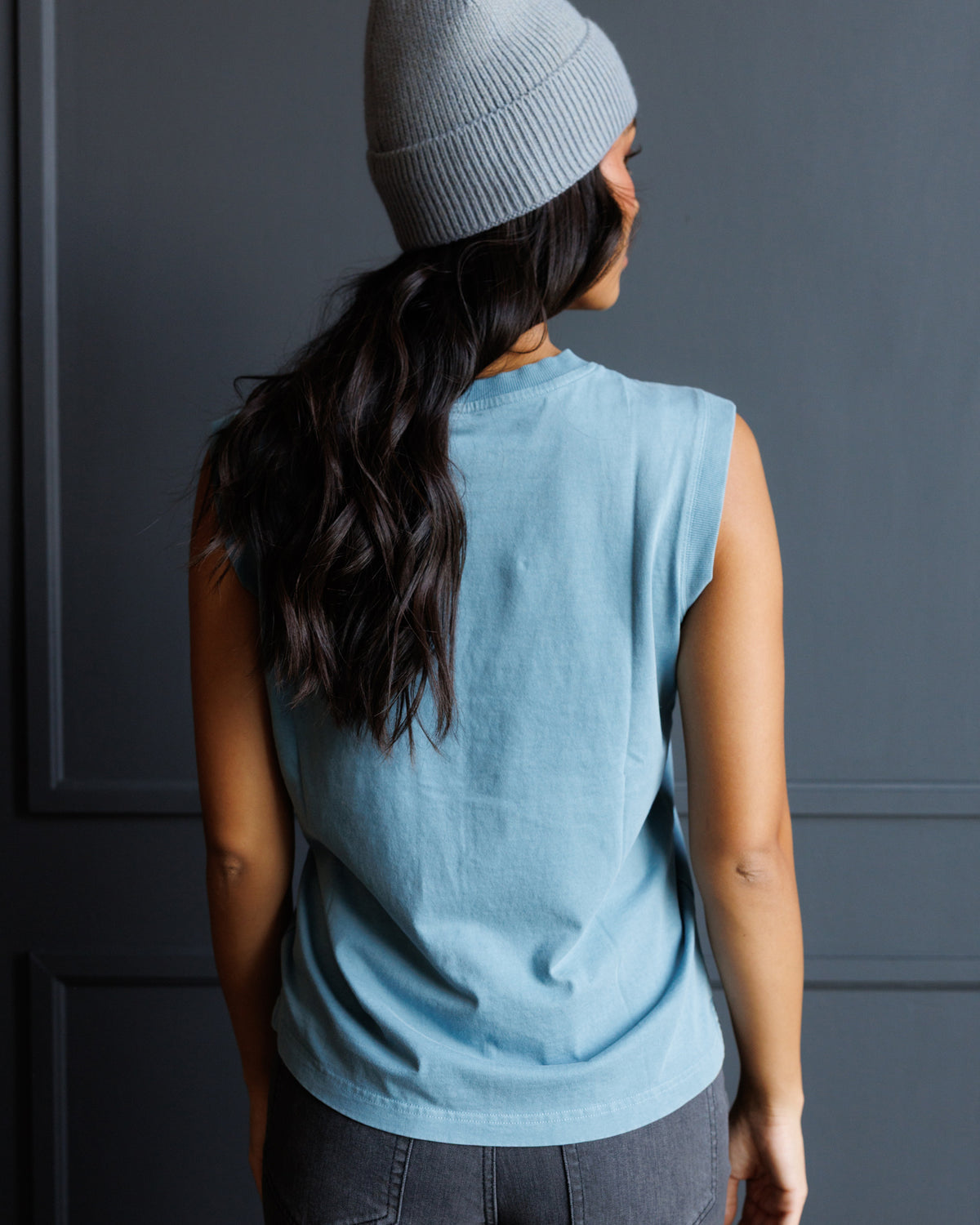 A woman shown from behind wearing a light blue sleeveless crop top, a light gray ribbed beanie, and dark gray pants, standing against a dark gray paneled wall.