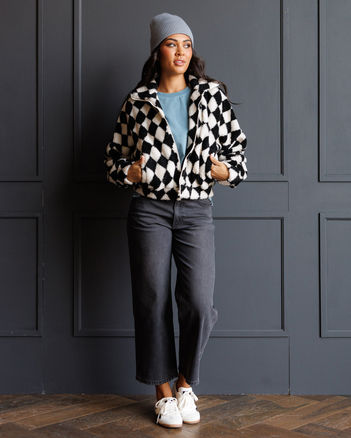 A woman stands against a dark paneled wall wearing a black and white checkered zip-up jacket, a light blue t-shirt, dark gray cropped jeans, white sneakers, and a gray beanie.