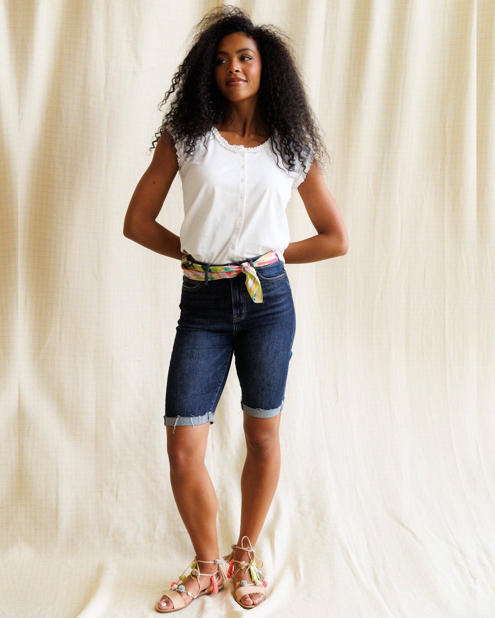 Woman wearing a white top and blue denim shorts with a colorful belt against a plain background