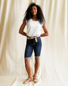 Woman wearing a white top and blue shorts with a colorful belt against a beige curtain background