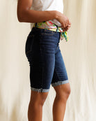 Person wearing dark blue denim shorts with a white shirt and colorful top underneath on a neutral background