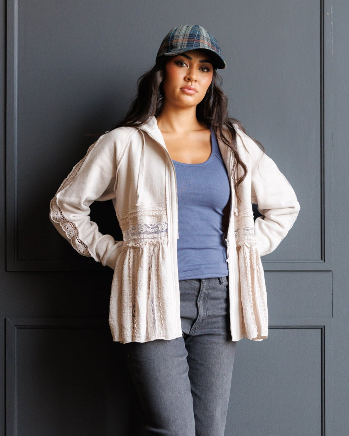 A woman stands against a dark gray paneled wall wearing a light beige hooded jacket over a slate blue top, gray jeans, and blue shoes, with a plaid cap on. The photo is taken in a real interior setting with wood flooring.