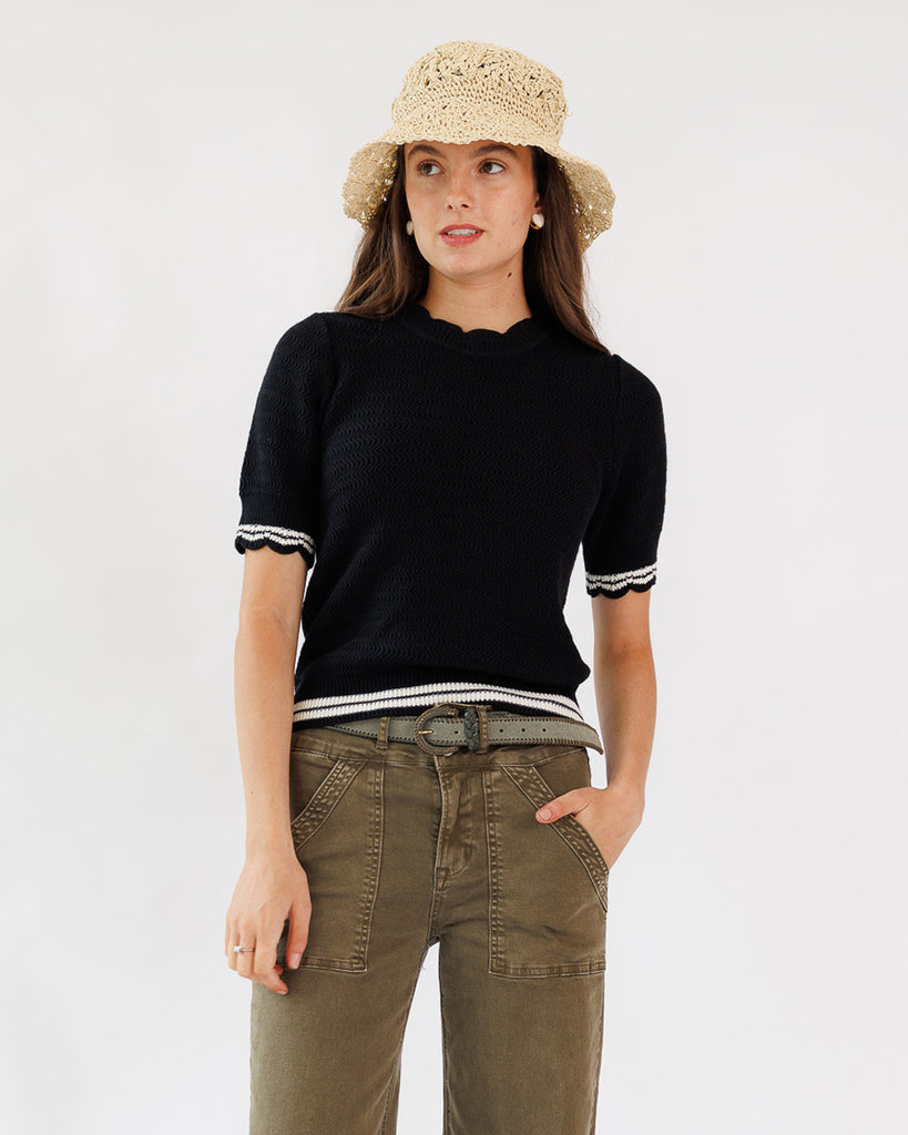 Woman wearing a black short-sleeve shirt with white trim, beige pants, and a straw hat on a light gray background
