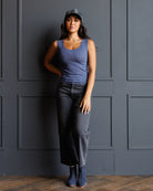 A woman wearing a slate blue tank top, dark gray cropped jeans, and a baseball cap leans against a dark gray paneled wall on a wooden floor.