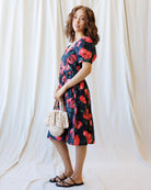 Woman wearing a floral dress holding a woven bag against a white curtain background