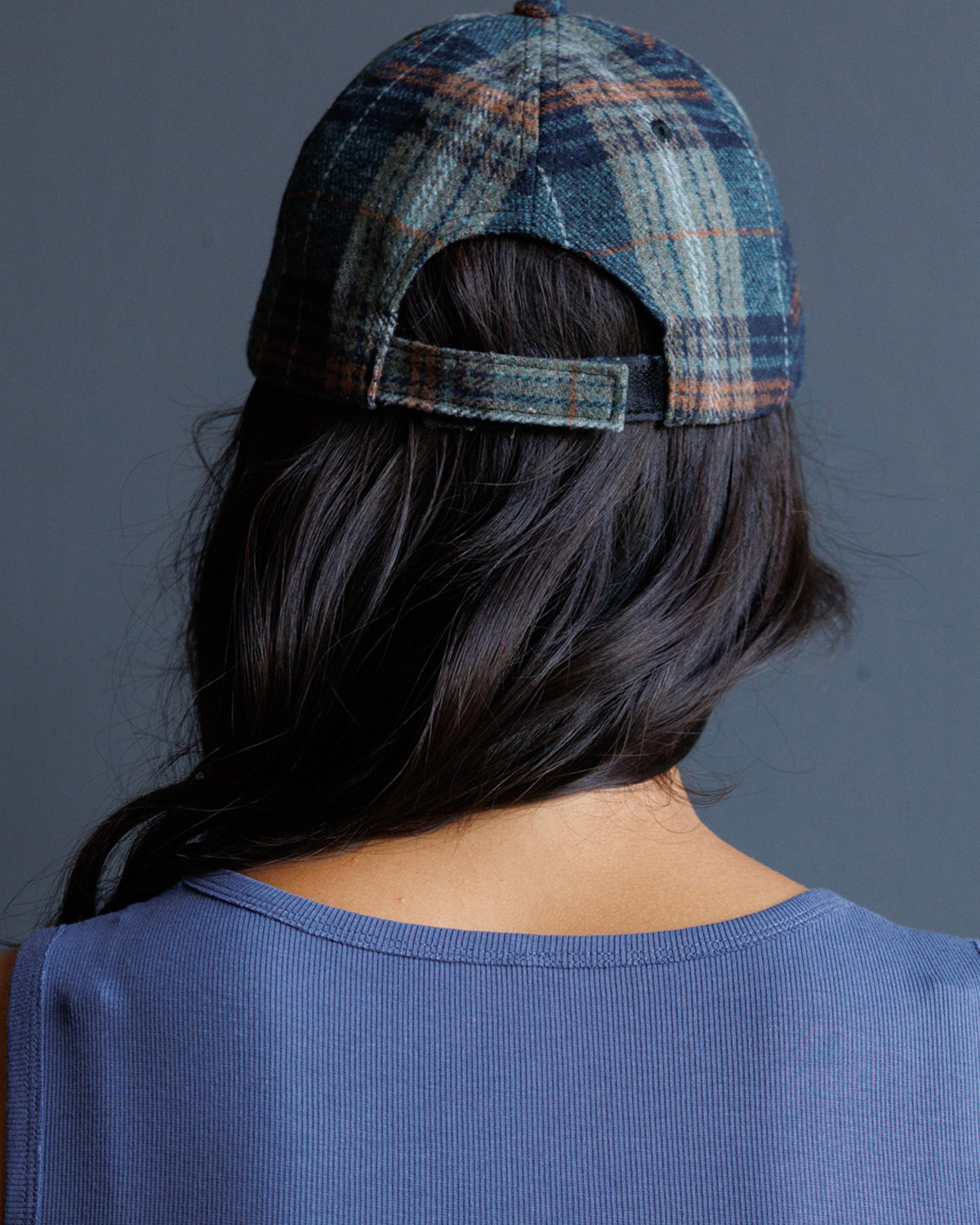 Rear view of a person wearing a blue and gray plaid baseball cap with a dark blue ribbed top against a dark gray background.