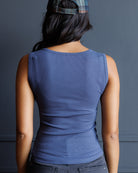 A woman viewed from behind wearing a fitted sleeveless blue top and a plaid cap against a dark gray wall.