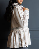 A woman wearing a cream-colored lace-trimmed cardigan, a plaid baseball cap, and dark gray pants, posing against a dark gray wall.