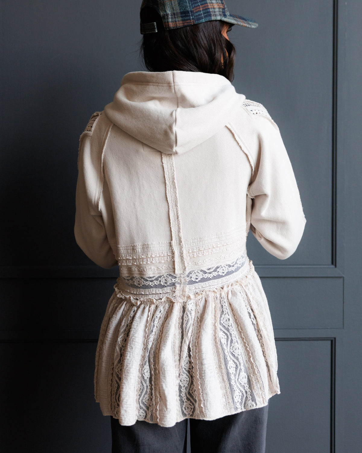 A person shown from behind wearing a cream-colored hooded jacket with lace detailing and a plaid cap, standing against a dark gray paneled wall.