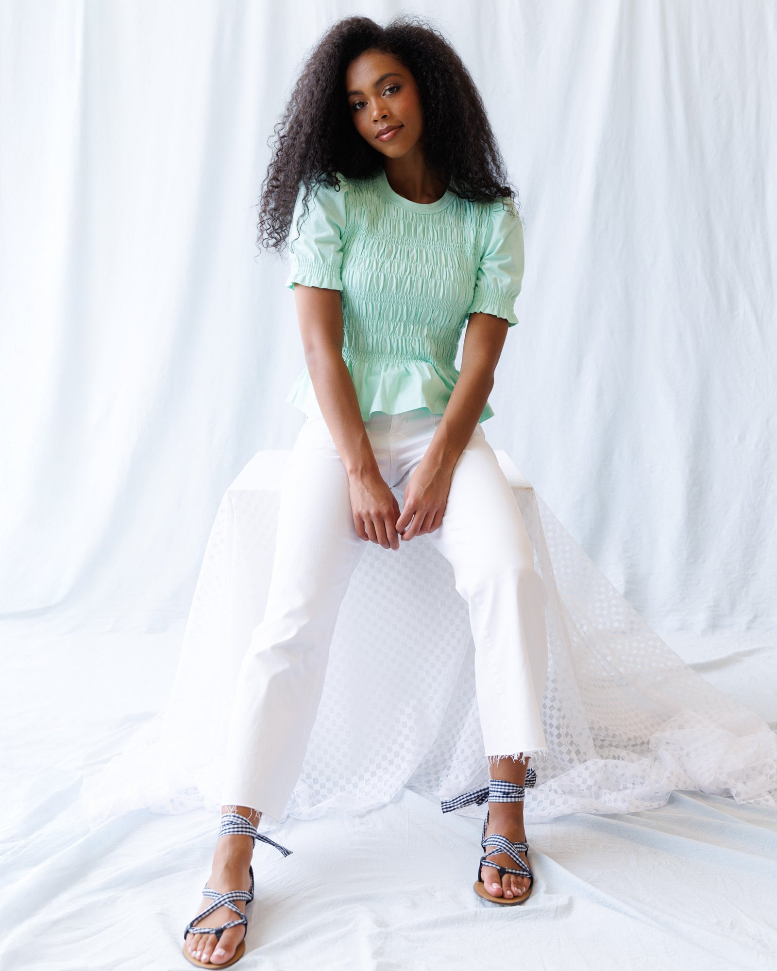Woman wearing a light green top and white pants sitting on a white fabric background