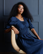 A woman with long, curly black hair wearing a navy blue midi dress with puff sleeves and a smocked waist, sitting on a light-colored wooden chair against a dark blue paneled wall.