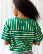 Person wearing a green and white striped shirt with a pocket, standing against a white curtain.