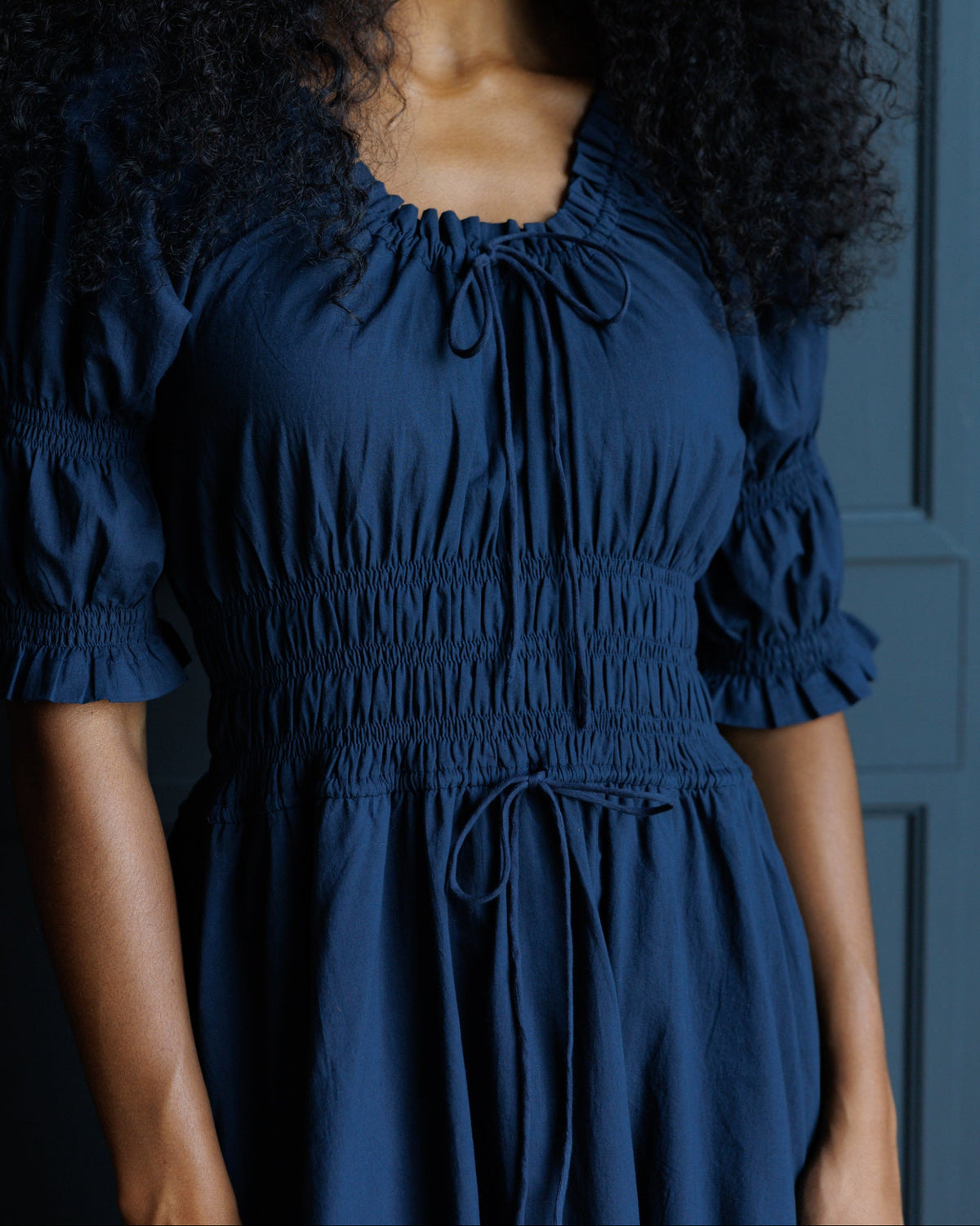 A woman wearing a dark blue smocked dress with puff sleeves and a drawstring neckline, shown from the shoulders to the hips against a dark blue paneled background.