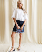 Woman wearing a white blouse and denim skirt with floral details against a plain background