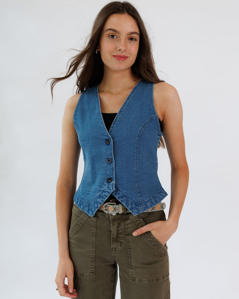 Woman wearing a blue denim vest over a black top and green pants on a white background