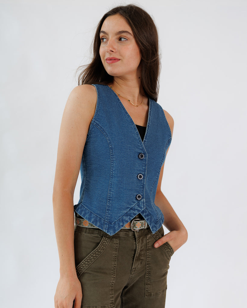 Woman wearing a blue denim vest over a black top and green pants on a white background