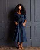 A woman with curly black hair stands against a dark gray paneled wall wearing a navy blue midi dress with ruffled sleeves and a gathered waist, paired with tan ankle boots.