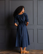 A woman stands with her back to the camera wearing a navy blue midi dress with puff sleeves and tan flat shoes, posing in front of a dark gray paneled wall.