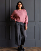 A woman stands against a dark gray paneled wall wearing a dusty rose cropped crewneck sweater, dark gray wide-leg jeans, and black lace-up sneakers.