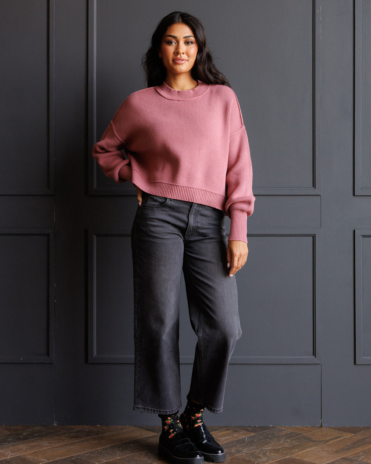 A woman stands against a dark gray paneled wall wearing a dusty rose cropped crewneck sweater, dark gray wide-leg jeans, and black lace-up sneakers.
