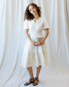 Woman wearing a white dress with a floral clutch against a white curtain background