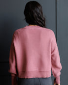 A woman viewed from behind wearing a dusty pink ribbed knit sweater and dark gray jeans, standing against a dark gray paneled wall.