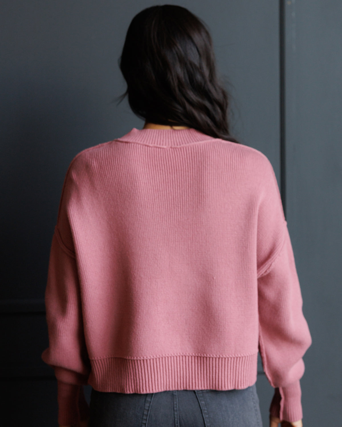 A woman viewed from behind wearing a dusty pink ribbed knit sweater and dark gray jeans, standing against a dark gray paneled wall.