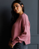 A woman wearing a dusty rose pink ribbed knit sweater and dark gray jeans, posing against a dark gray paneled wall.
