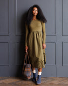 A woman stands against a dark gray paneled wall wearing an olive green long-sleeve midi dress with a gathered waist and eyelet lace hem. She is holding a plaid tote bag and wearing blue ankle boots and white socks.
