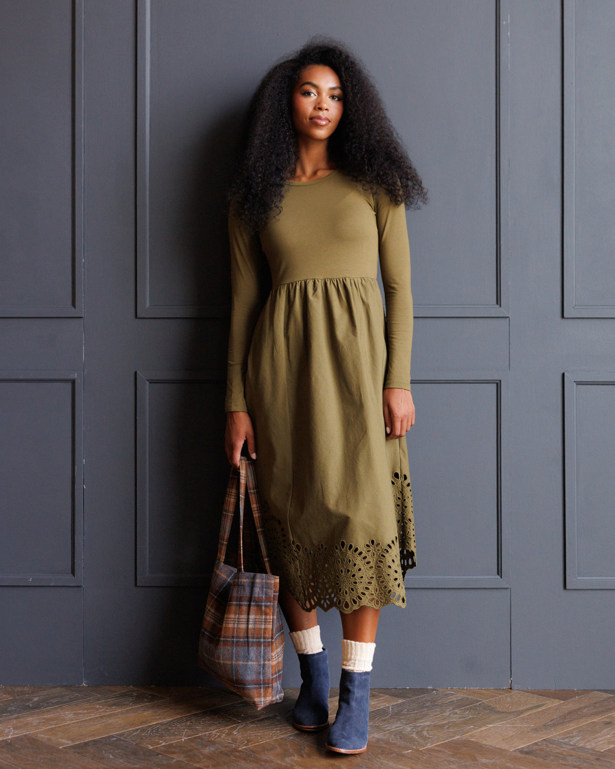 A woman stands against a dark gray paneled wall wearing an olive green long-sleeve midi dress with a gathered waist and eyelet lace hem. She is holding a plaid tote bag and wearing blue ankle boots and white socks.