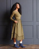 A woman wearing a long-sleeve olive green midi dress with a scalloped eyelet hem, paired with white socks and blue suede boots, standing against a dark gray paneled wall.