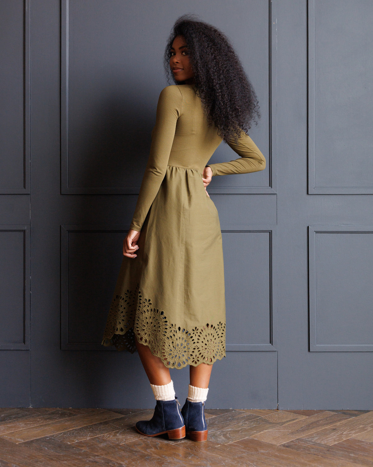 A woman with long curly dark hair is shown from the back wearing an olive green long-sleeve midi dress with a scalloped eyelet hem. She is standing in front of a dark gray paneled wall on a wooden floor.