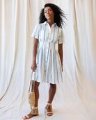 Woman wearing a striped dress holding a woven bag against a white curtain background