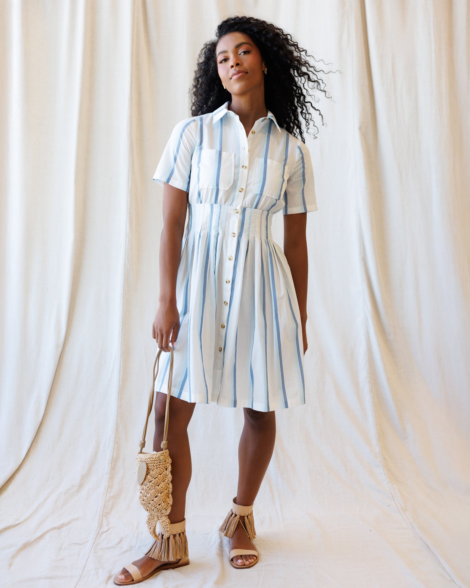 Woman wearing a striped dress holding a woven bag against a white curtain background