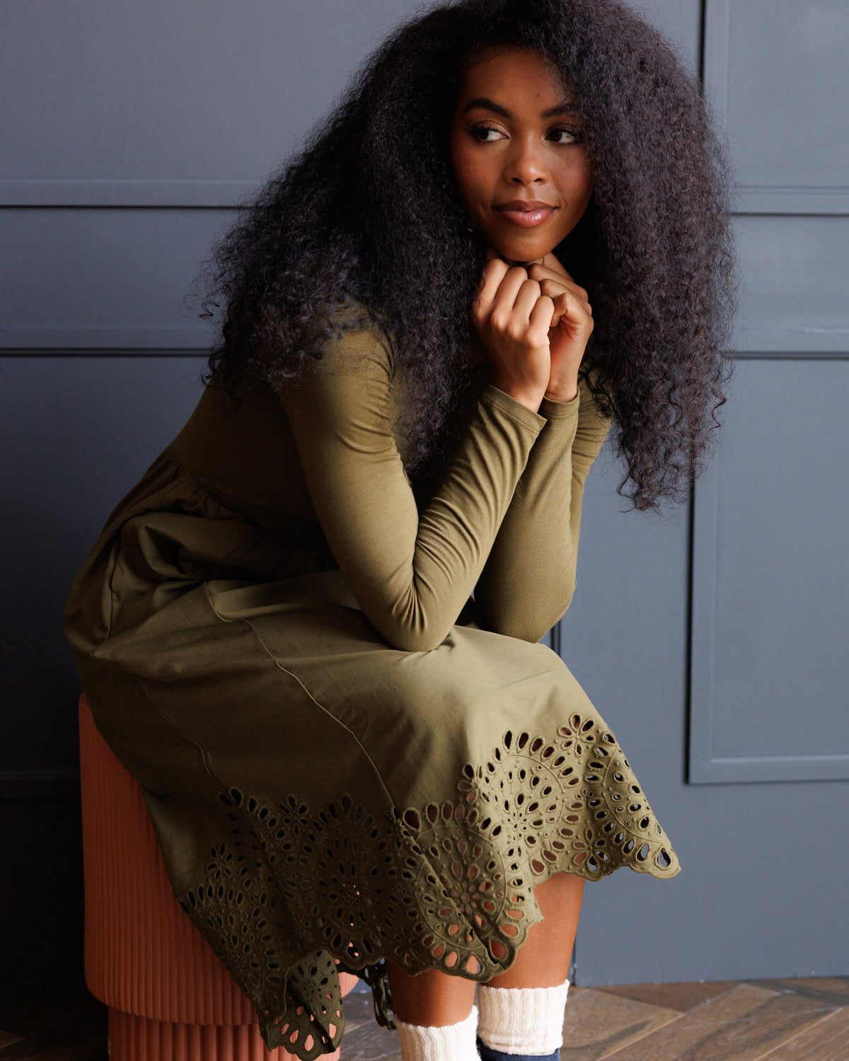 A woman with long, curly black hair sits on a terracotta stool wearing an olive green long-sleeve dress with a scalloped eyelet hem. She is posed against a dark gray paneled wall.