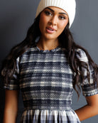 A woman wearing a white knit beanie and a smocked short-sleeve dress with a navy, white, and gray plaid skirt, posed against a dark gray studio background.