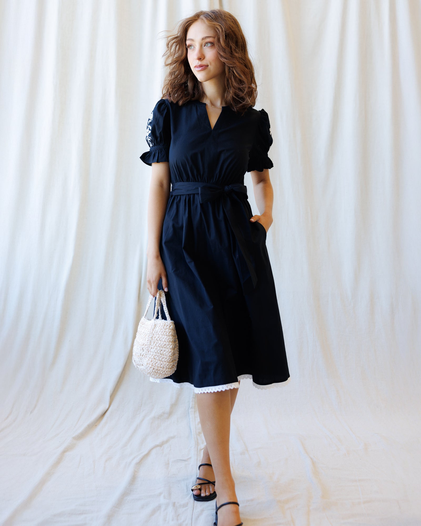 Woman wearing a black dress holding a white handbag against a white curtain background
