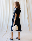 Woman wearing a black dress with white embroidery, holding a woven bag against a white curtain background.