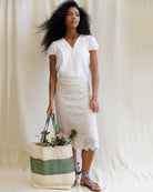 Woman holding a woven bag with flowers, wearing a white blouse and beige lace skirt against a plain background